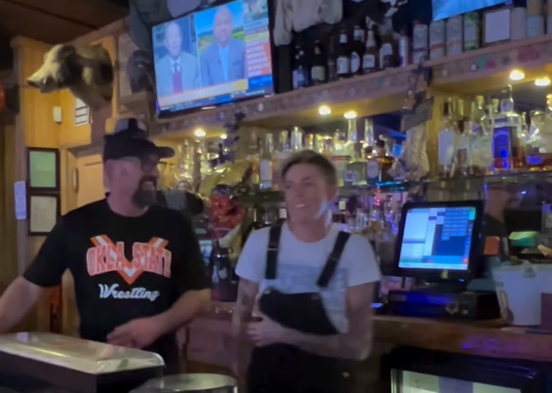Behind the bar at the Rawhide on a December night: bottles ranked two shelves deep under the screens, shaded lamps along the counter, and a bartender in overalls laughing