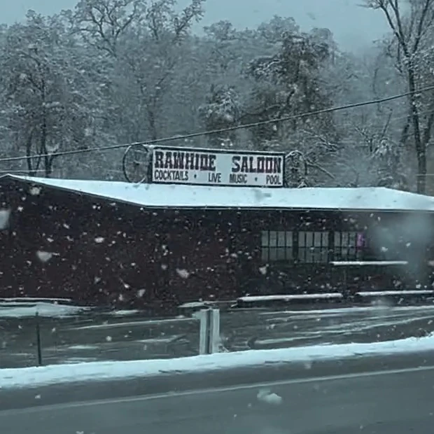 The Rawhide Saloon from Highway 108 in the snow: the sign board on its wagon-wheel brackets reading RAWHIDE SALOON, with COCKTAILS, LIVE MUSIC and POOL beneath it, over the long dark roadhouse and its snow-covered roof