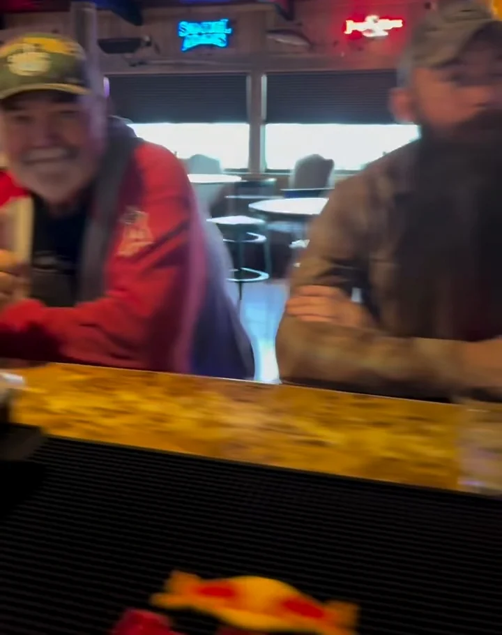 Two regulars laughing at the bar rail under the neon beer signs, the daylight windows and the pool room behind them